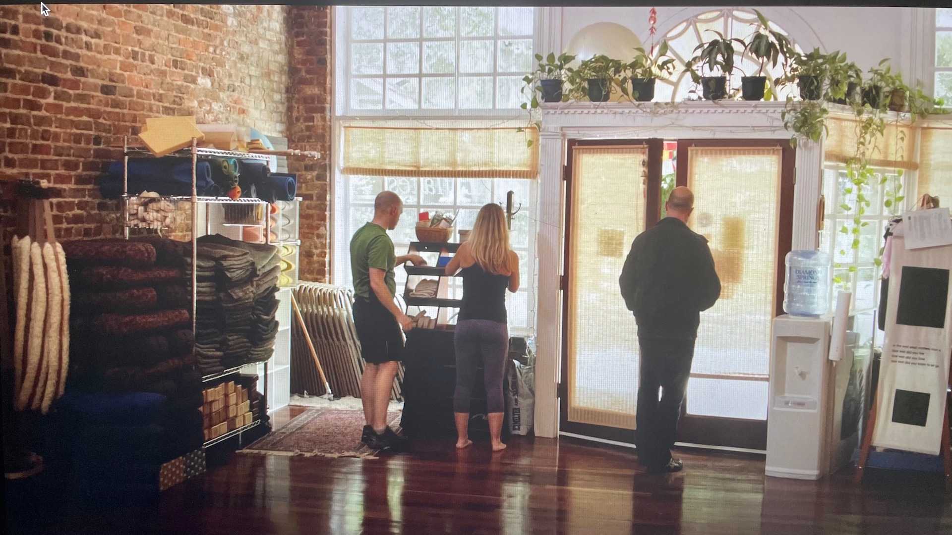 The inside of 8th Street Yoga Studio facing toward 8th Street, sunlight streaming in. Bolsters, Blankets, and Blocks stacked on the left against the brick wall. To the right Scott Lundgren in a green t-shirt, gray shorts, blonde woman to the right arranging face towels on a bookcase in front of a window. A slim bald man in a dark green jacket and black pants leaving the studio.